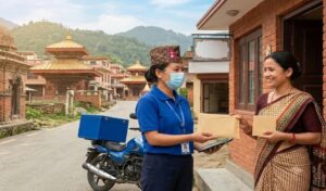 नेपालको ग्रामीण क्षेत्रमा एक पोशाक लगाएकी पोस्टल डेलिभरी कर्मचारीले ब्रिक भवन बाहिर परम्परागत पहिरनमा रहेकी महिलालाई एउटा खाम हस्तान्तरण गरिरहेकी छन्, पृष्ठभूमिमा मन्दिर र हरिया डाँडाकाँडा देखिन्छन्।